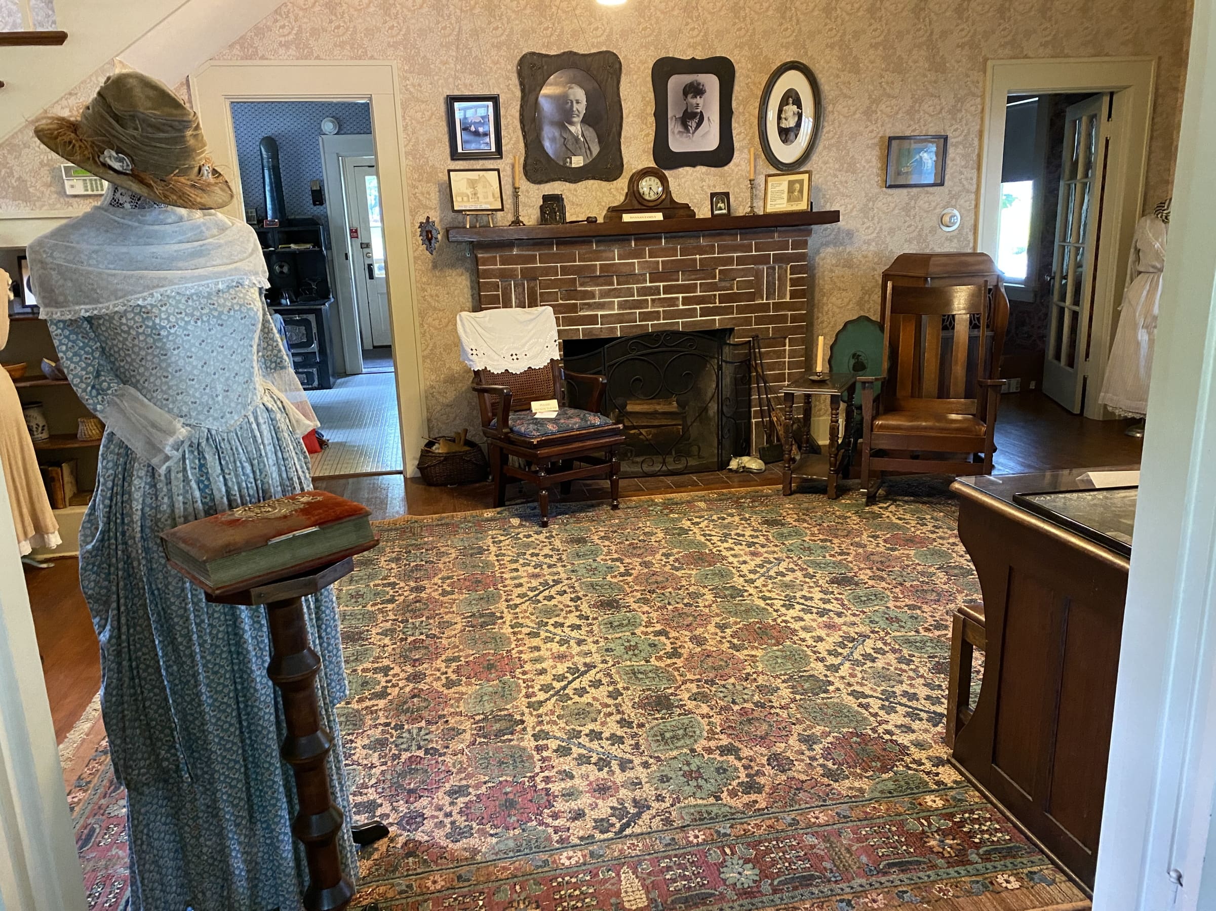 Inside of the Bothell Historical Museum. The room looks like an ol living room and has a fireplace with old photos on the wall. To the left of the image, there is a dressform wearing a very old blue dress. The room has been kept in tact to look like its the 1820s.