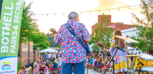 Photo taken from on stage behind the subjects. An older man and younger woman play guitar on stage to a group at a Summer Nights in Bothell event.